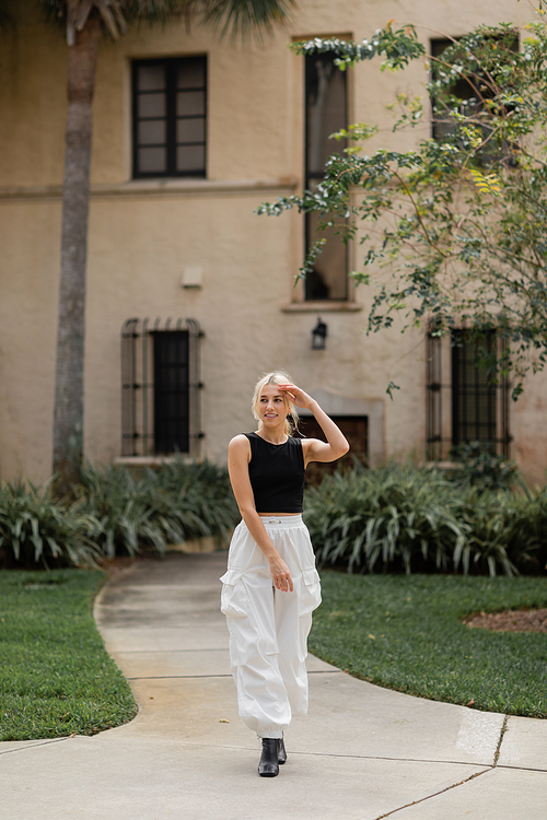 full length of pleased young woman in white cargo pants and trendy tank top standing near house in Miami