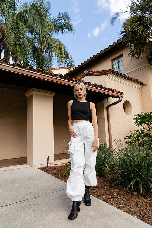 full length of young woman in trendy clothes and boots standing near modern house and green plants in Miami