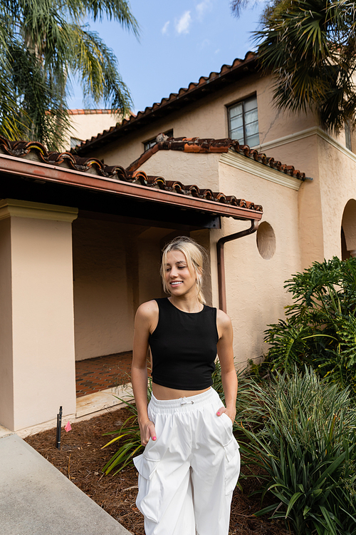 happy young woman in trendy clothes standing with hands in pockets near modern house in Miami