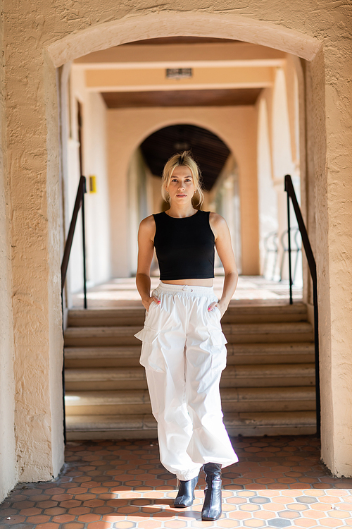 full length of blonde woman in stylish outfit standing with hands in pockets near stairs in modern house in Miami