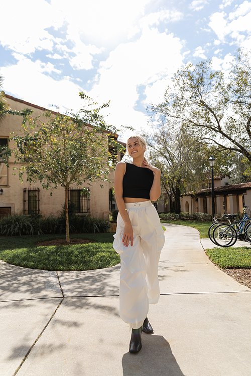 full length of positive woman in trendy clothes standing in stylish boots near modern house in Miami