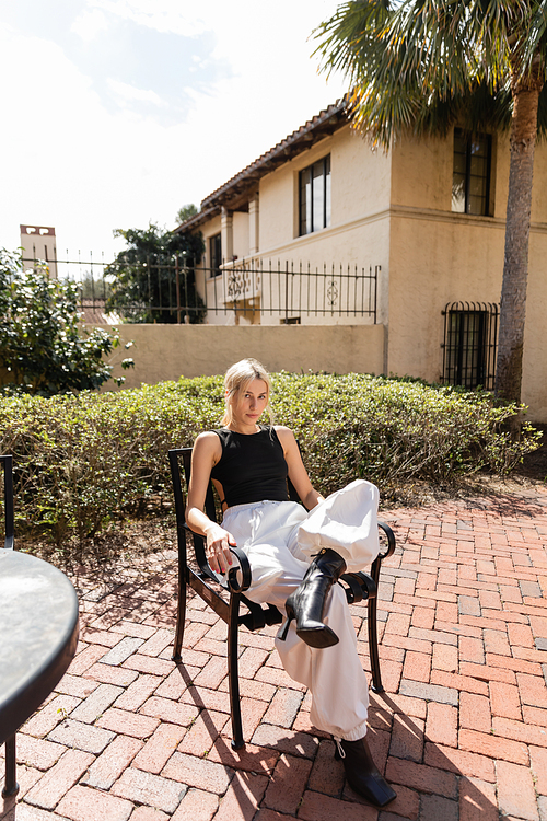 full length of young woman in trendy clothes sitting on patio chair near modern house in Miami