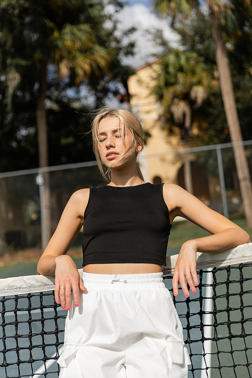 young woman in black tank top and white cargo pants leaning on net in tennis court