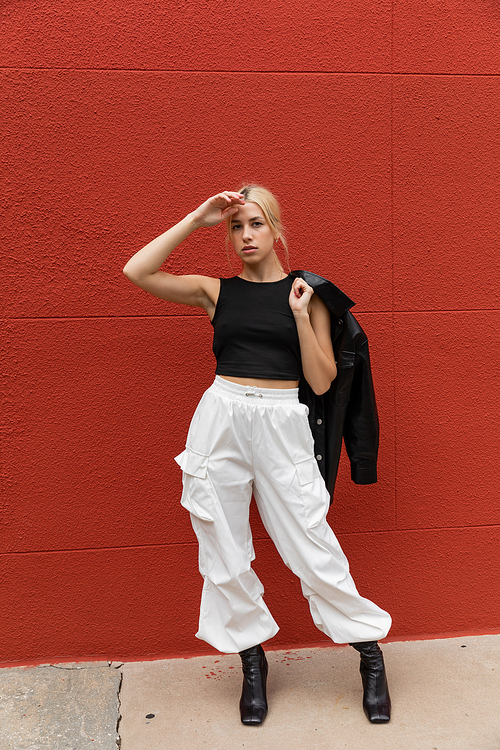 full length of stylish woman in cargo pants standing with leather shirt jacket near red wall in Miami