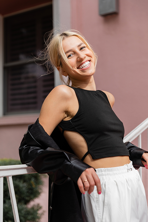cheerful blonde woman in black tank top and cargo pants standing near pink house in Miami