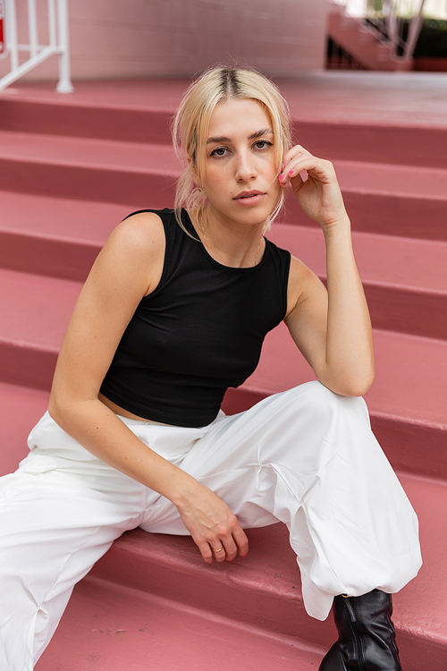 blonde young woman in black tank top and cargo pants sitting on pink stairs in Miami
