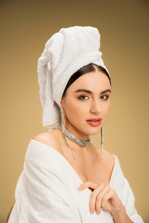 young woman in jewelry with white towel on head looking at camera on beige background