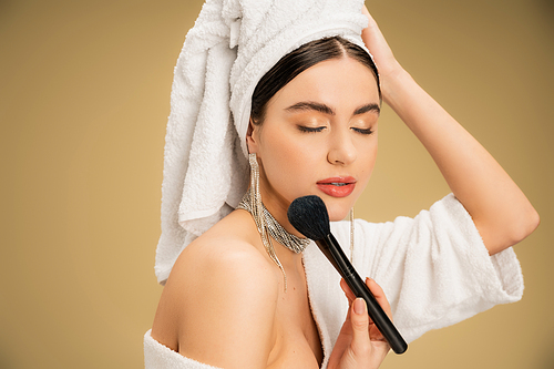 brunette woman with white towel on head applying face powder with makeup brush on beige background