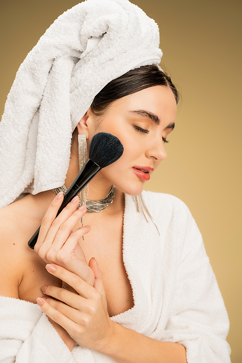 young woman with white towel on head applying face powder with makeup brush on beige background