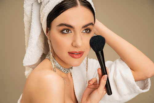 brunette woman with white towel on head applying face powder with makeup brush on grey background