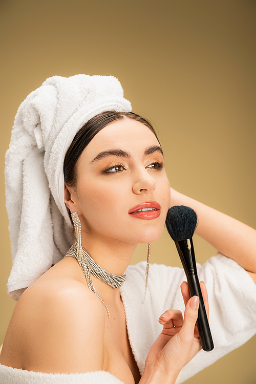 charming woman in white towel on head applying face powder with makeup brush on beige background