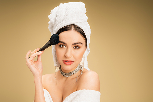 charming woman with white towel on head applying face powder with makeup brush on beige background