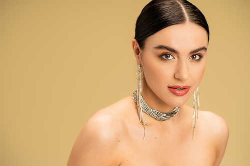 brunette young woman in necklace and earrings looking at camera isolated on beige