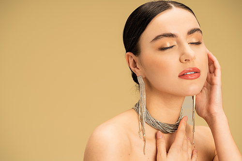 brunette young woman in necklace and earrings posing with closed eyes isolated on beige