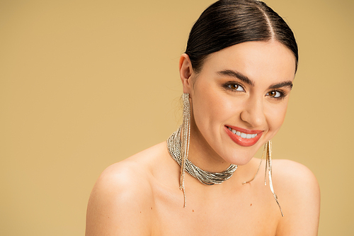 joyful woman in necklace and earrings looking at camera isolated on beige