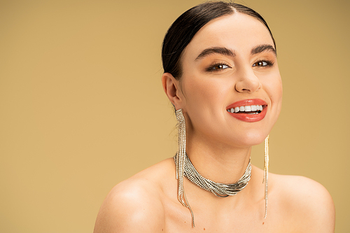 elegant woman in necklace and earrings looking at camera while smiling isolated on beige