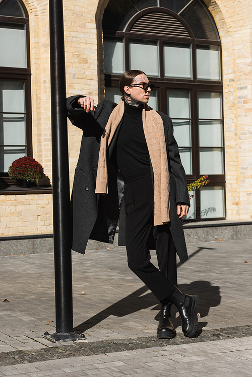 full length of tattooed man in trendy coat and stylish sunglasses standing with crossed legs on urban street