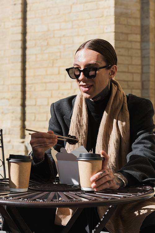 stylish tattooed man in sunglasses and coat holding chopsticks while eating asian food
