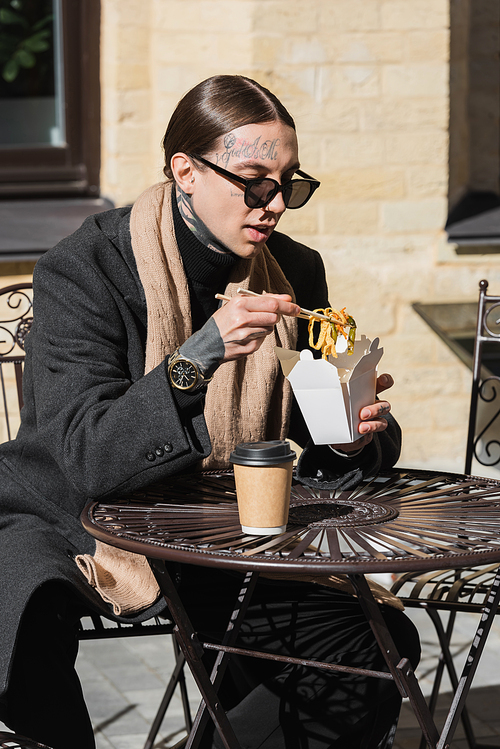 tattooed man in stylish sunglasses holding chopsticks while eating asian food near paper cup on bistro table