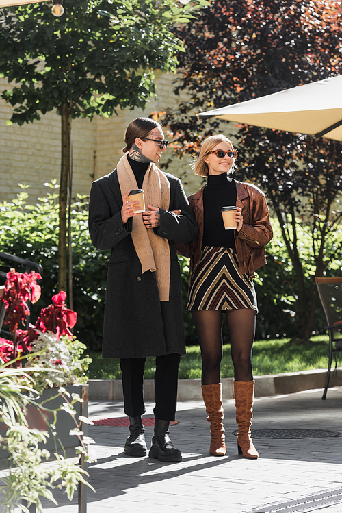 full length of stylish couple in sunglasses holding paper cups while standing near outdoor cafe