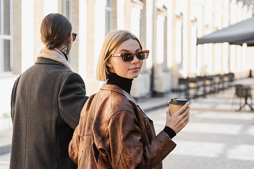 blonde woman in trendy sunglasses holding paper cup while walking with man during date
