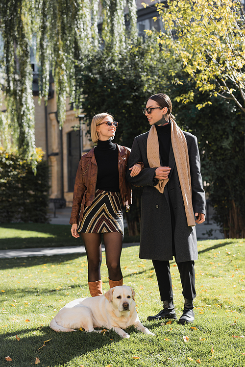 full length of cheerful couple in sunglasses walking with labrador in park