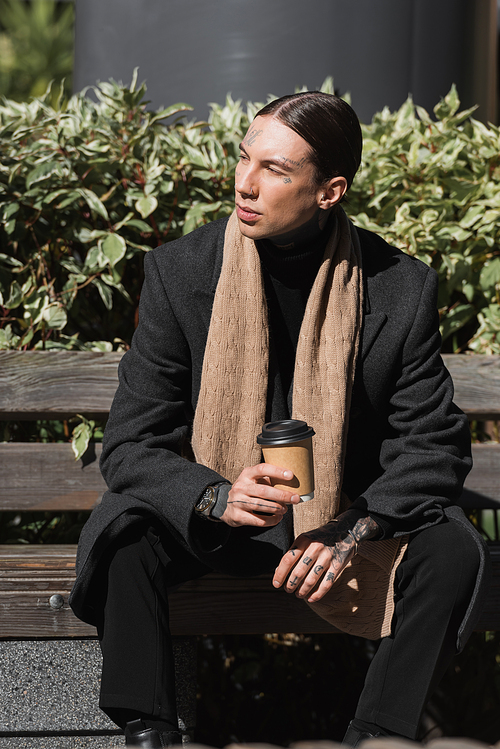 young tattooed man in coat and scarf holding takeaway drink while sitting on bench