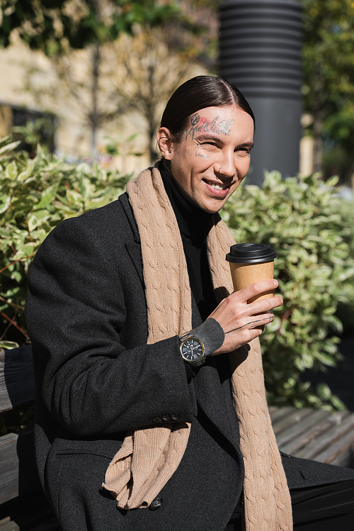happy tattooed man in coat and scarf holding takeaway drink while sitting on bench