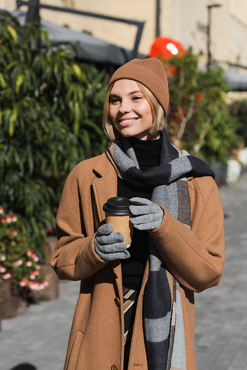 pleased woman in stylish coat and beige hat holding paper cup with coffee to go