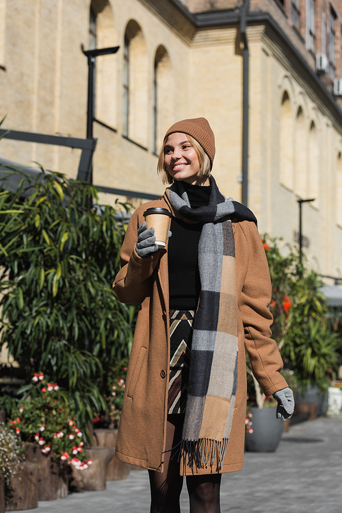 smiling young woman in stylish coat and beige hat holding paper cup with coffee to go