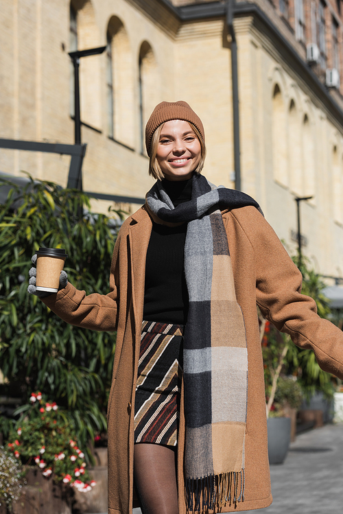 happy woman in stylish coat and beige hat holding paper cup with coffee to go