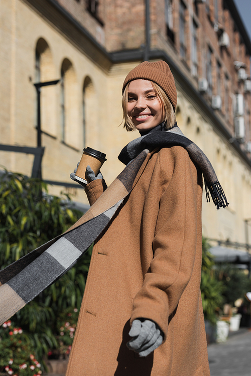 joyful young woman in beige coat and hat holding paper cup with coffee to go