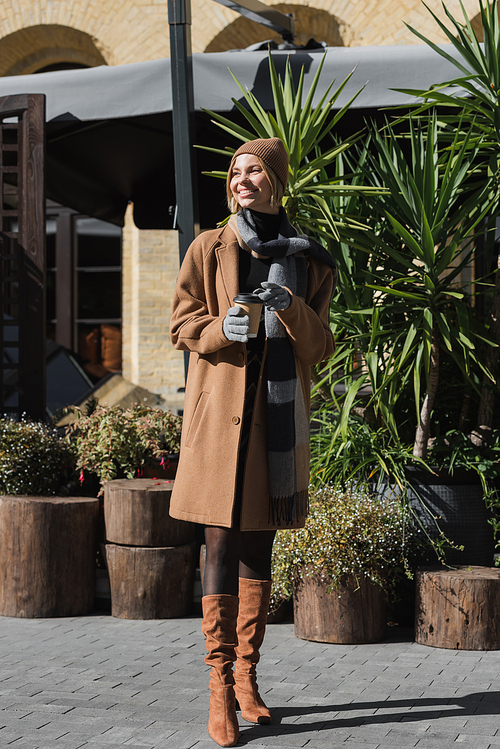 full length of happy blonde woman in beige coat and hat holding paper cup with coffee to go