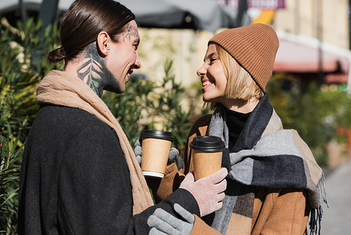 side view of happy blonde woman in beige hat looking at tattooed boyfriend in coat while holding coffee to go