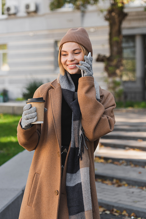 happy blonde woman in hat and coat holding paper cup while talking on smartphone