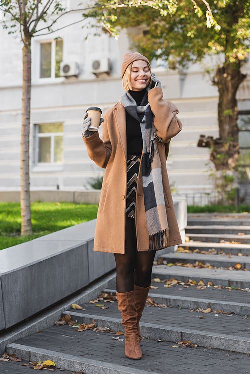 full length of happy blonde woman in hat and coat holding paper cup while talking on smartphone
