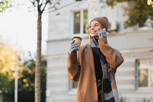 smiling woman in hat and coat holding paper cup while talking on smartphone