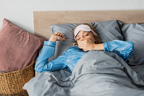 sleepy woman in silk pajama and night mask yawning and covering mouth while lying in bed