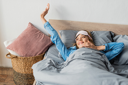 cheerful woman in sleeping mask and nightwear stretching while lying in bed