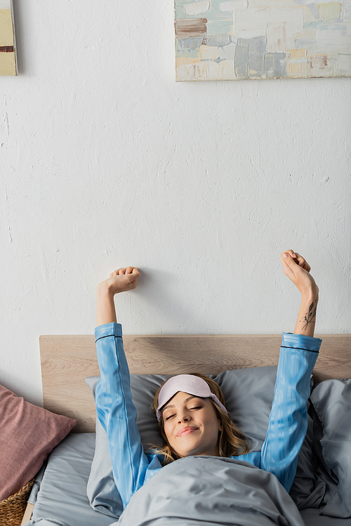 tattooed woman in sleeping mask and nightwear stretching while lying in bed