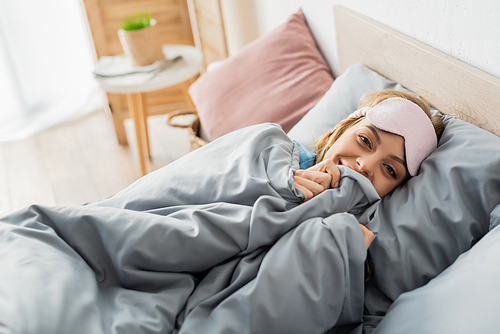 happy young woman in sleeping mask lying under blanket in comfortable bed