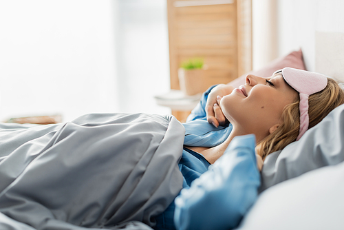 side view of pleased woman in sleeping mask lying under blanket in comfortable bed