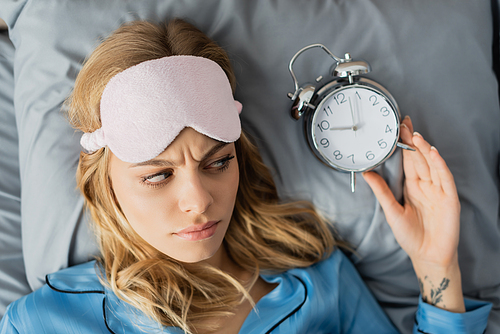 top view of tattooed woman in sleeping mask and blue pajama looking at alarm clock while lying in bed