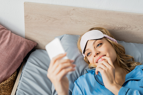 high angle view of cheerful woman in sleeping mask and blue pajama using smartphone in bed