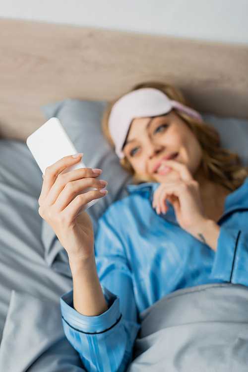 blurred and cheerful woman in sleeping mask and blue pajama using smartphone in bed