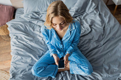 top view of cheerful woman in blue pajama using smartphone with blank screen while sitting on bed