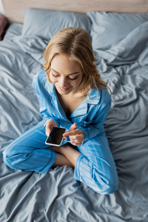 top view of smiling woman in blue pajama using smartphone with blank screen while sitting on bed