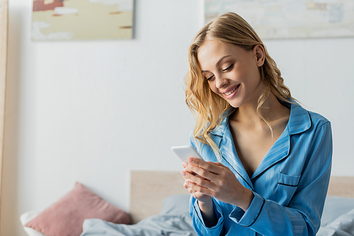 pleased woman in blue pajama using mobile phone in bedroom