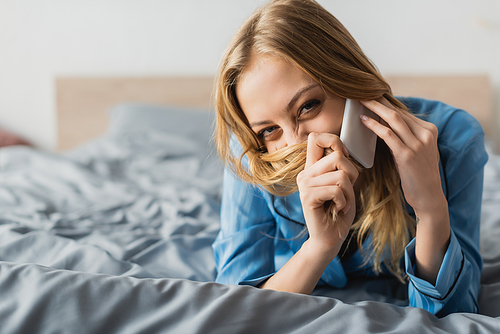 cheerful woman talking on smartphone while covering face with blonde hair