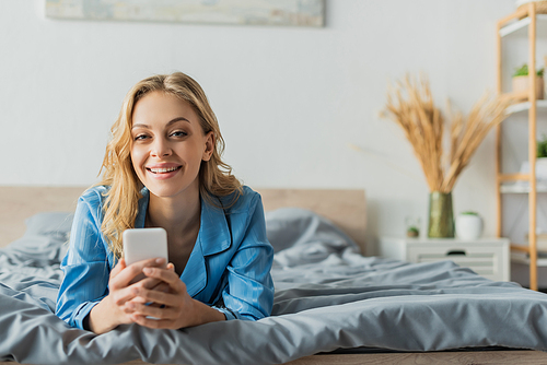 joyful young woman messaging on smartphone while lying in blue pajama on bed